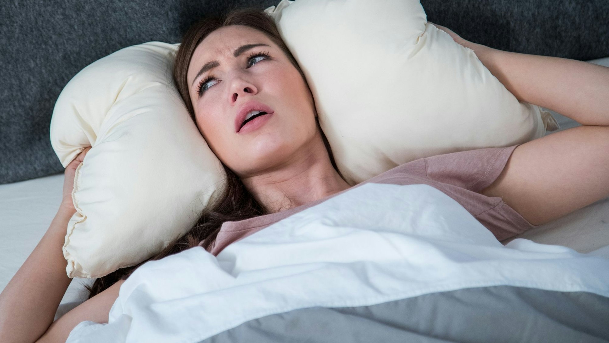 Genervte Frau liegt im Bett und drückt ihr Kissen mit ihren Händen rechts und links auf ihre Ohren.
