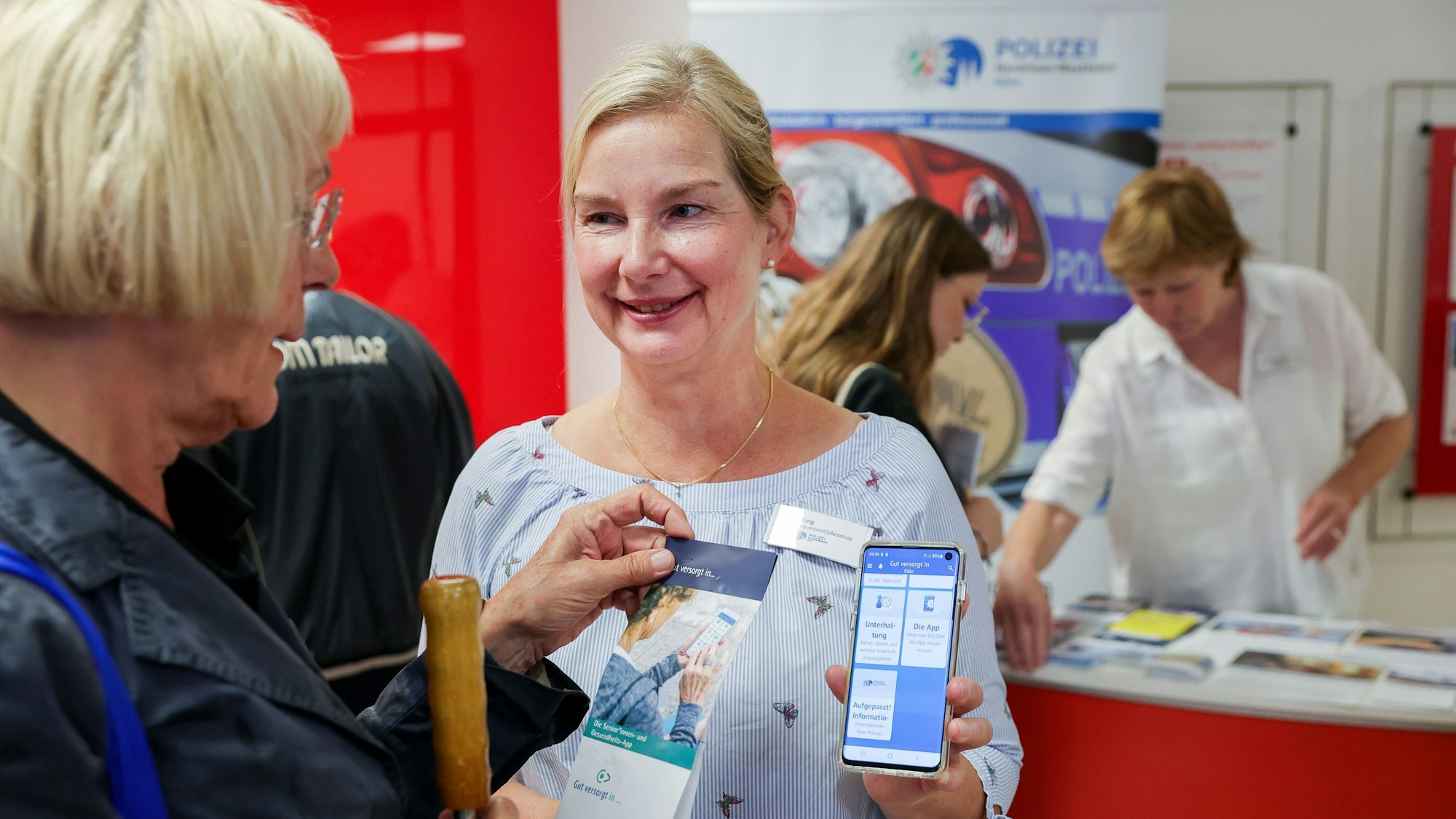 Polizistin Sigrid Jung erklärt einer Seniorin die App „Gut versorgt in“. Sie hält ein Handy mit der geöffneten App. Die Seniorin hält einen Flyer.