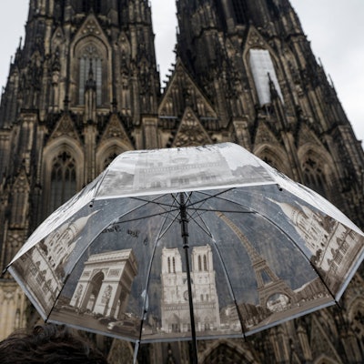 Eine Touristin hält am Kölner Dom einen Regenschirm mit Sehenswürdigkeiten, um sich vor dem Regen zu schützen. Nordrhein-Westfalen hat im Vergleich der Bundesländer im Juli 2023 die wenigsten Sonnenstunden erlebt. (Symbolbild)