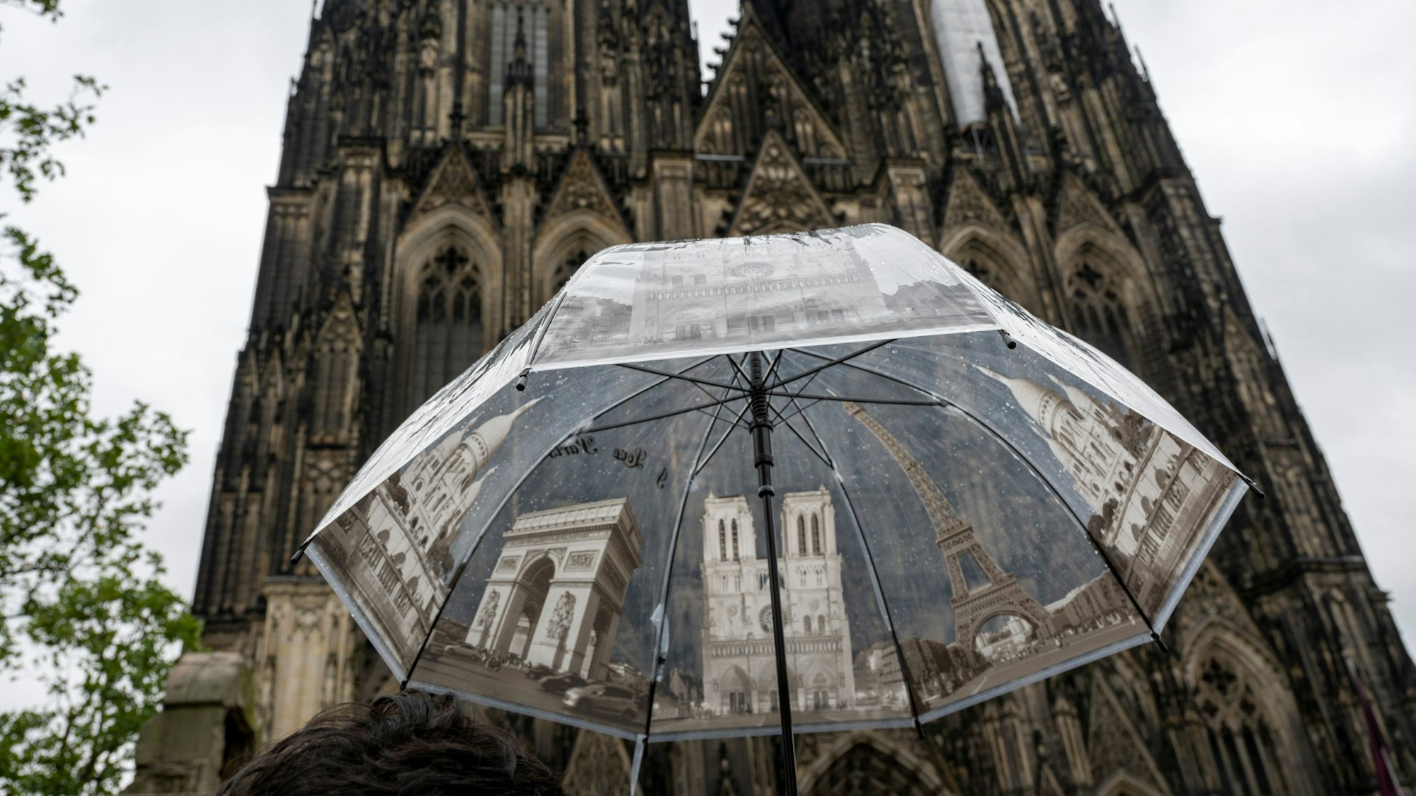 Eine Touristin hält am Kölner Dom einen Regenschirm mit Sehenswürdigkeiten, um sich vor dem Regen zu schützen. Nordrhein-Westfalen hat im Vergleich der Bundesländer im Juli 2023 die wenigsten Sonnenstunden erlebt. (Symbolbild)