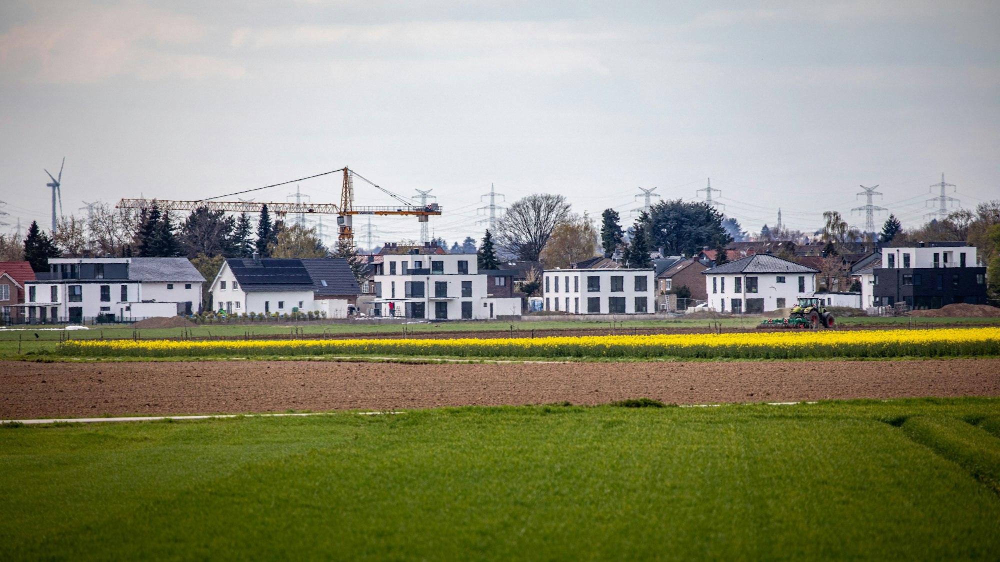 Neu errichtete Wohnhäuser stehen am Ortsrand, im Hintergrund ist ein Baukran zu sehen. Vor den Einfamilienhäusern breiten sich Ackerflächen und grüne Wiesen aus.