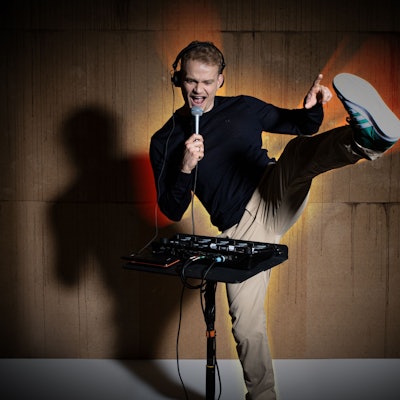 Das Bild zeigt den Künstler Dustin Waree hinter einem Mischpult. In der Hand hält er ein Mikrofon.