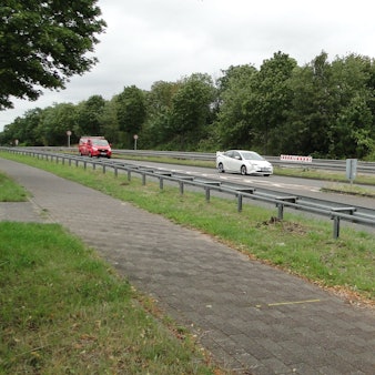 Autos fahren über eine Landstraße, ein Radweg ist durch Leitplanken abgetrennt.