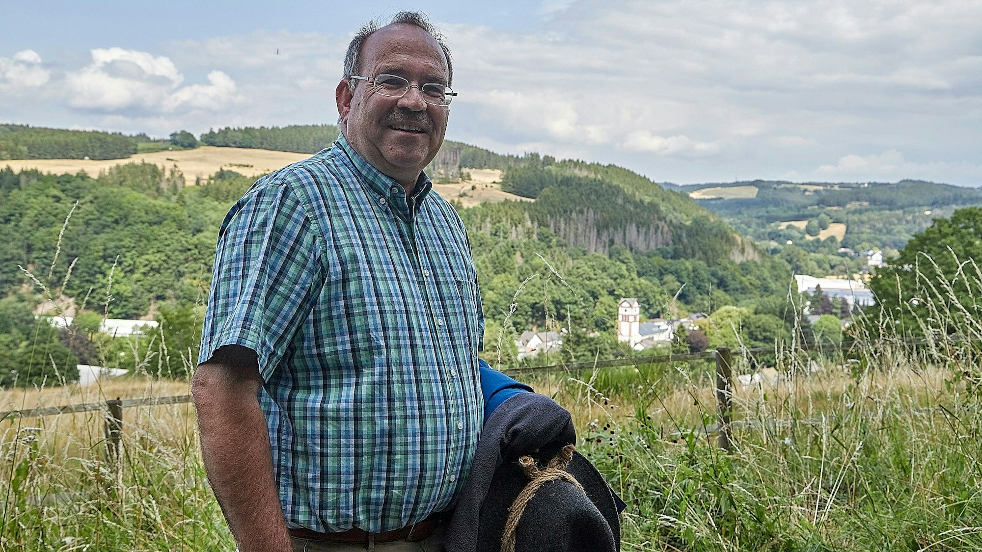 Ein Mann im rustikalen Wanderoutfit steht vor einer sommerlichen Eifellandschaft.
