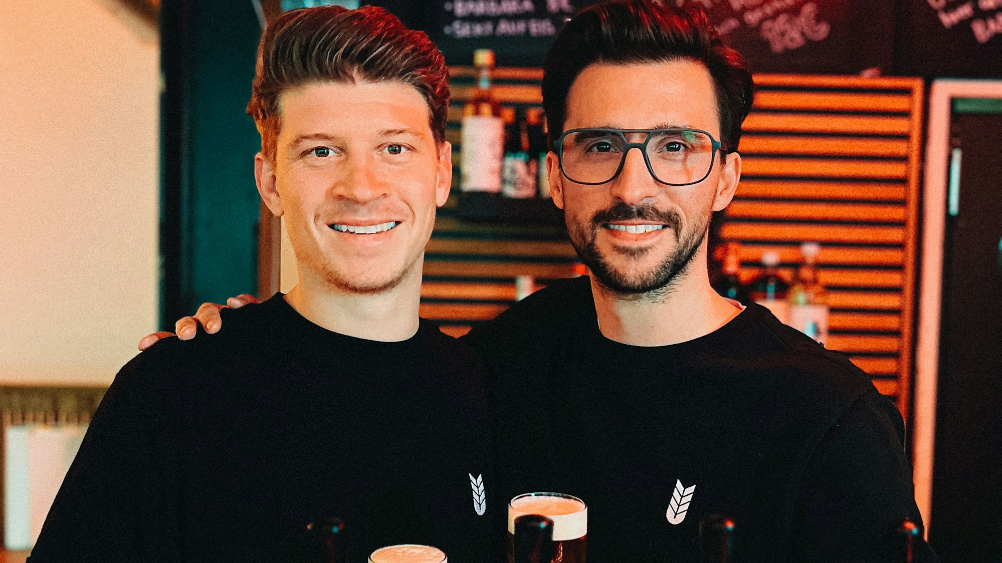 Die Gründer Nicolas Lutz (l.) und Maximilian Koeser (r.) in ihrer heimischen „Brennerbude“. Beide halten ein Glas Zappes-Pils in der Hand.