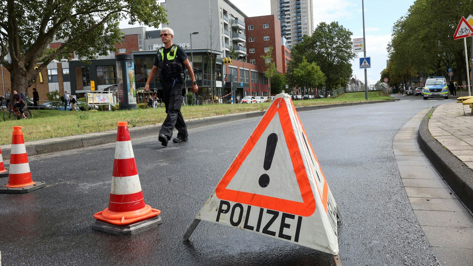Ein Polizist geht auf der gesperrten, leeren Straße entlang. Im Vordergrund steht ein Warndreieck mit der Aufschrift Polizei.