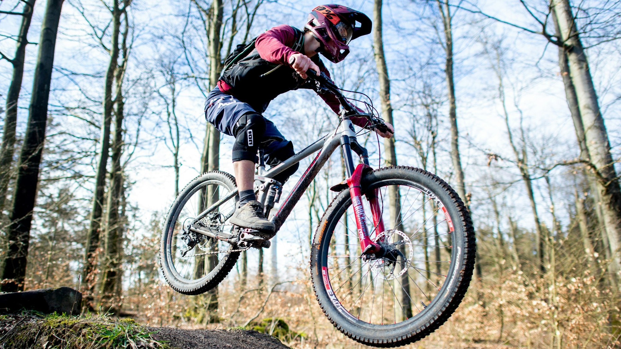 Ein junger Mann springt mit seinem Mountainbike über eine Anhöhe auf einer Downhillstrecke.
