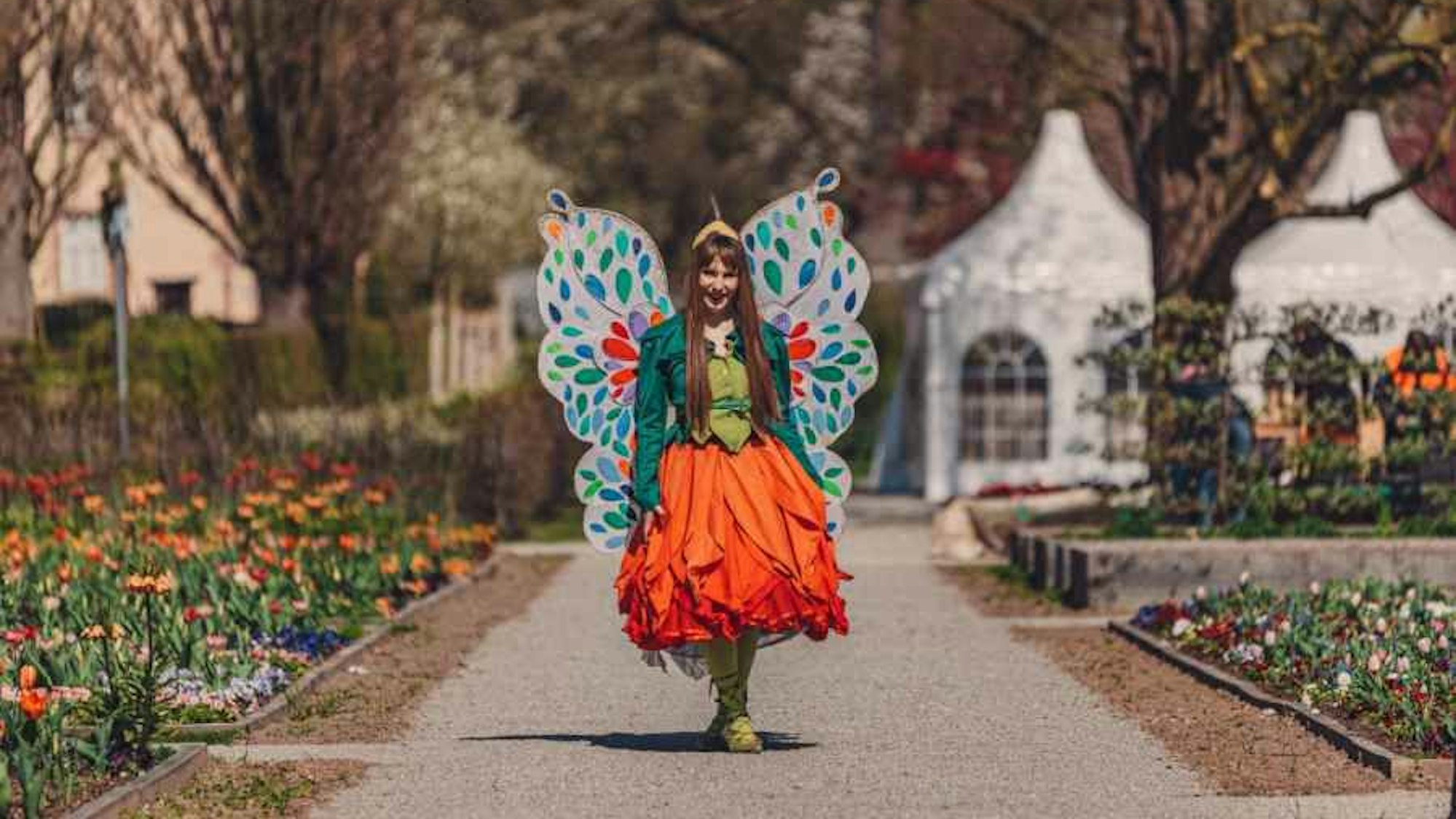 Eine Frau spaziert in einem Feen-Kostüm mit großen Flügeln und orangenem Rock über einen Weg zwischen Blumenbeeten.