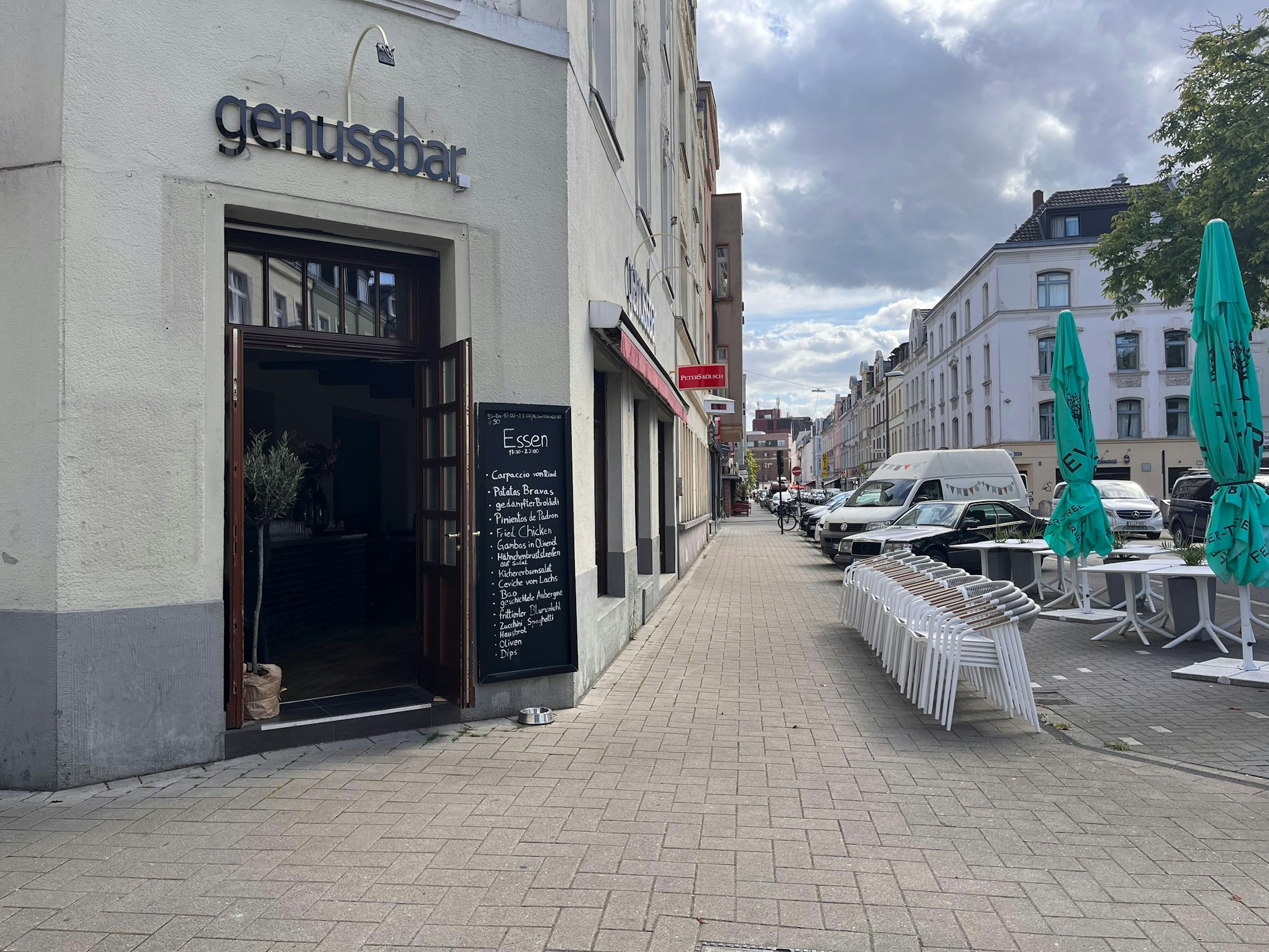 Über einem Ecklokal ist das Schild Genussbar zu sehen. An der Straße stehen Stühle und Tische.