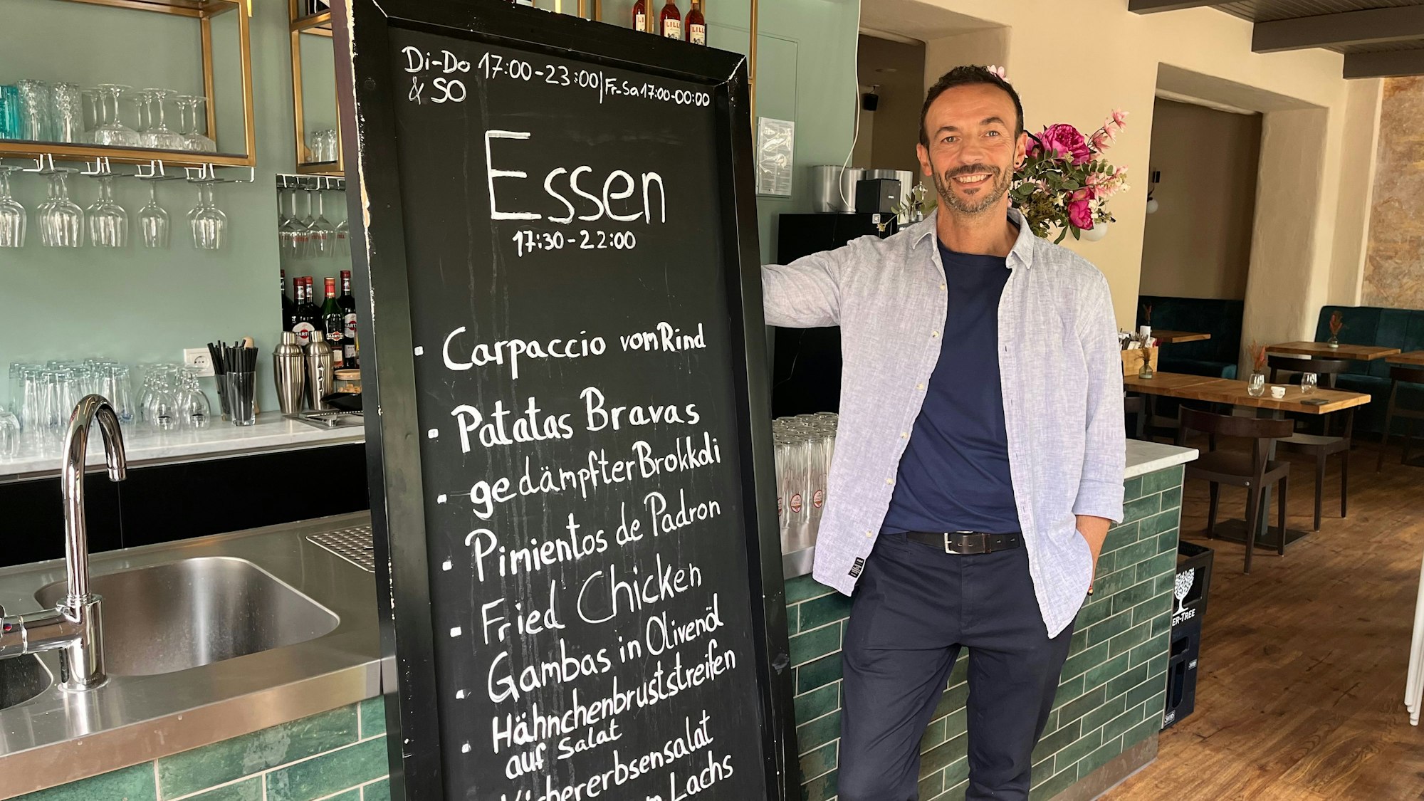 Ein junger Mann steht neben einer großen Tafel, auf der Gerichte angeschrieben sind, vor einem grün gefliesten Tresen in einem Restaurant.