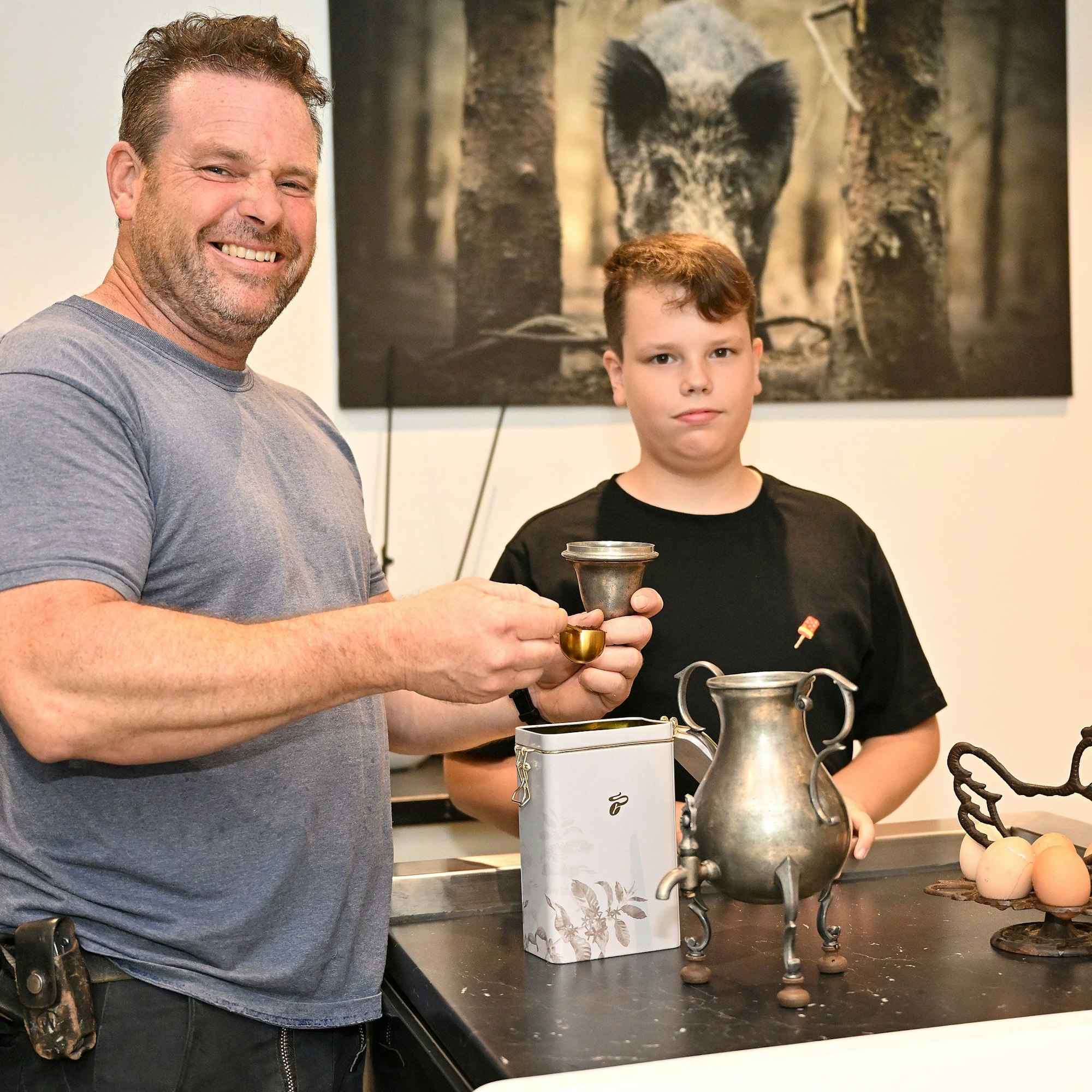 Ein Vater steht mit seinem Sohn in der Küche und füllt Kaffeepulver in den Dekel einer Zinnkanne.