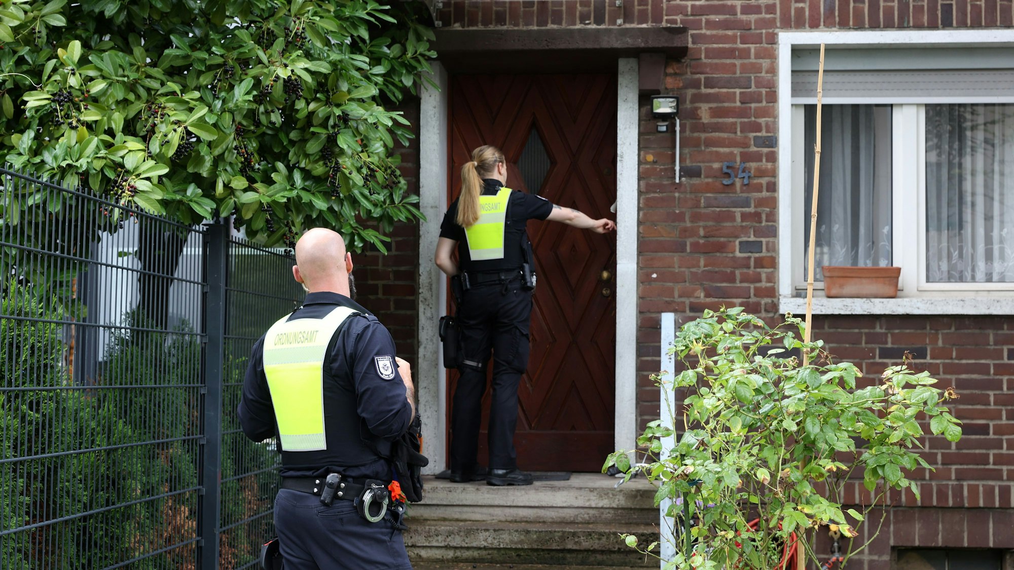 Eine Frau in Einsatzbekleidung klingelt an einem Wohnhaus. Im Vordergrund steht ihr Kollege. Ihre Uniformen sind teilweise in greller Warnfarbe gehalten.