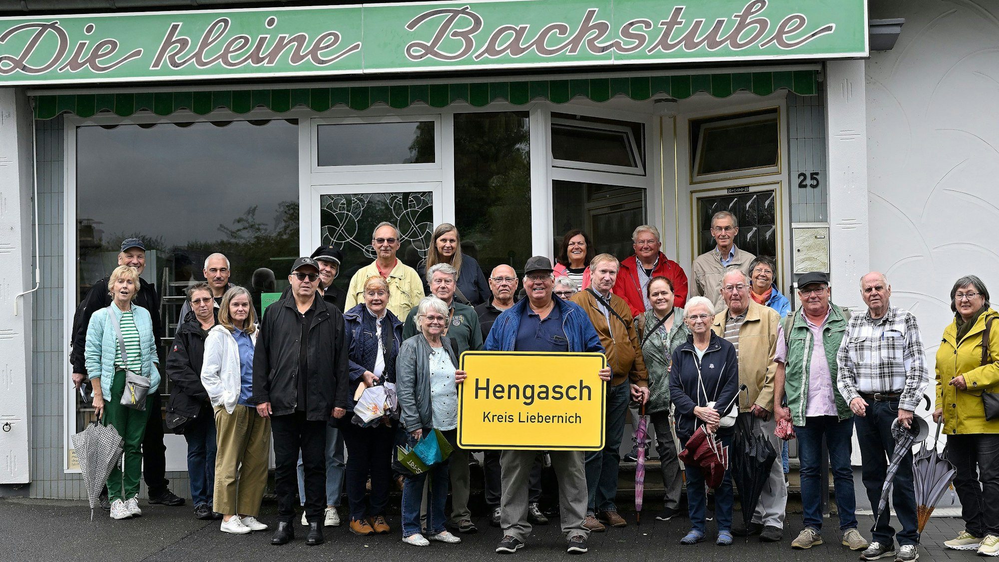 Menschen stehen mit einer gelben Ortstafel, auf der zu lesen ist „Hengasch Kreis Liebernich“ vor einem Ladenlokal mit der Aufschrift „Die kleine Backstube“.