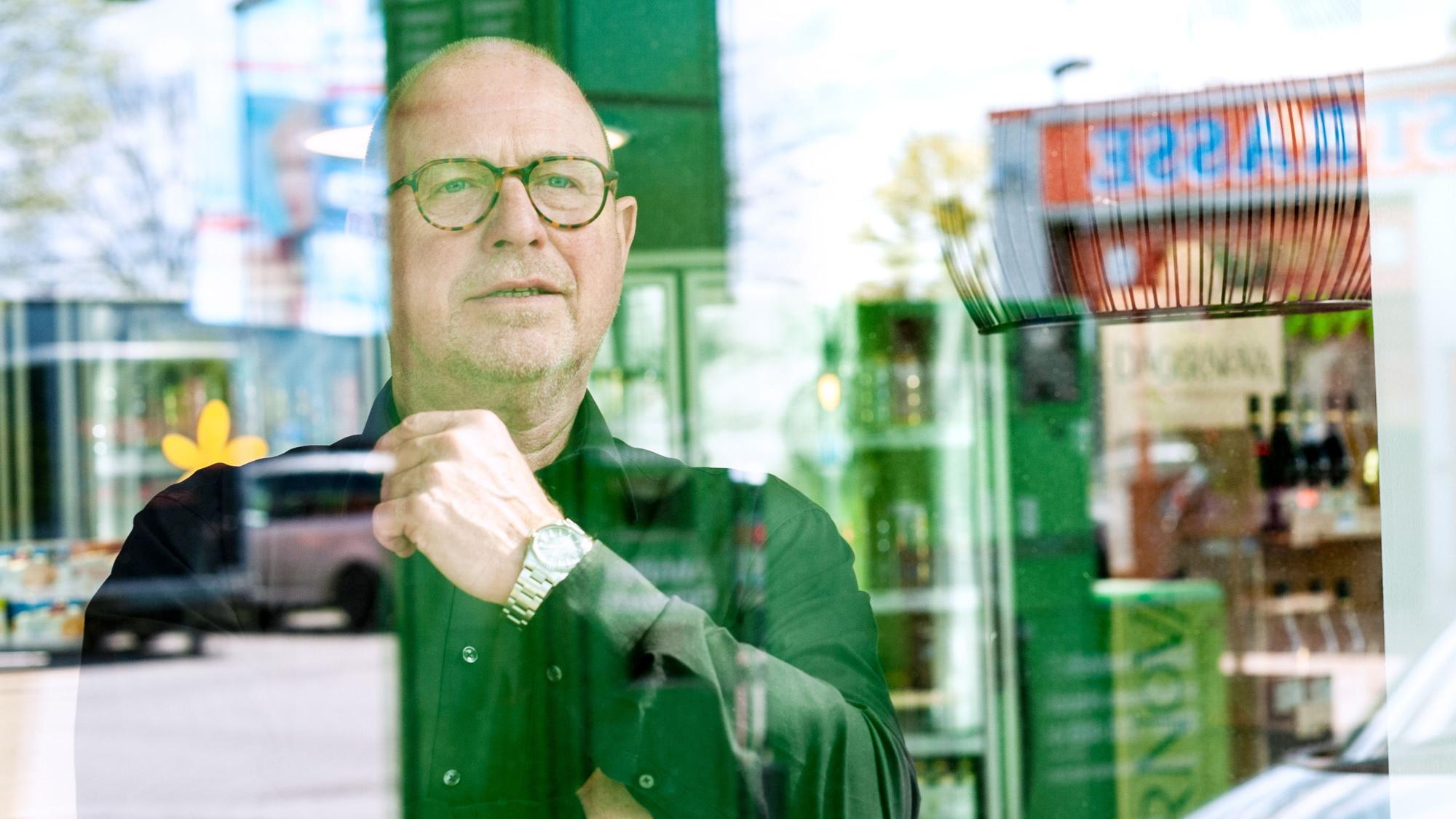Karl-Heinz Breuer schaut aus dem Schaufenster, in dessen Spiegelung Teile der Tankstelle zu sehen sind. Hinter ihm stehen Getränke.