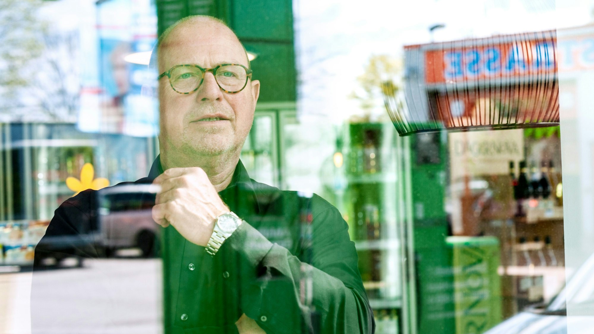 Karl-Heinz Breuer schaut aus dem Schaufenster, in dessen Spiegelung Teile der Tankstelle zu sehen sind. Hinter ihm stehen Getränke.