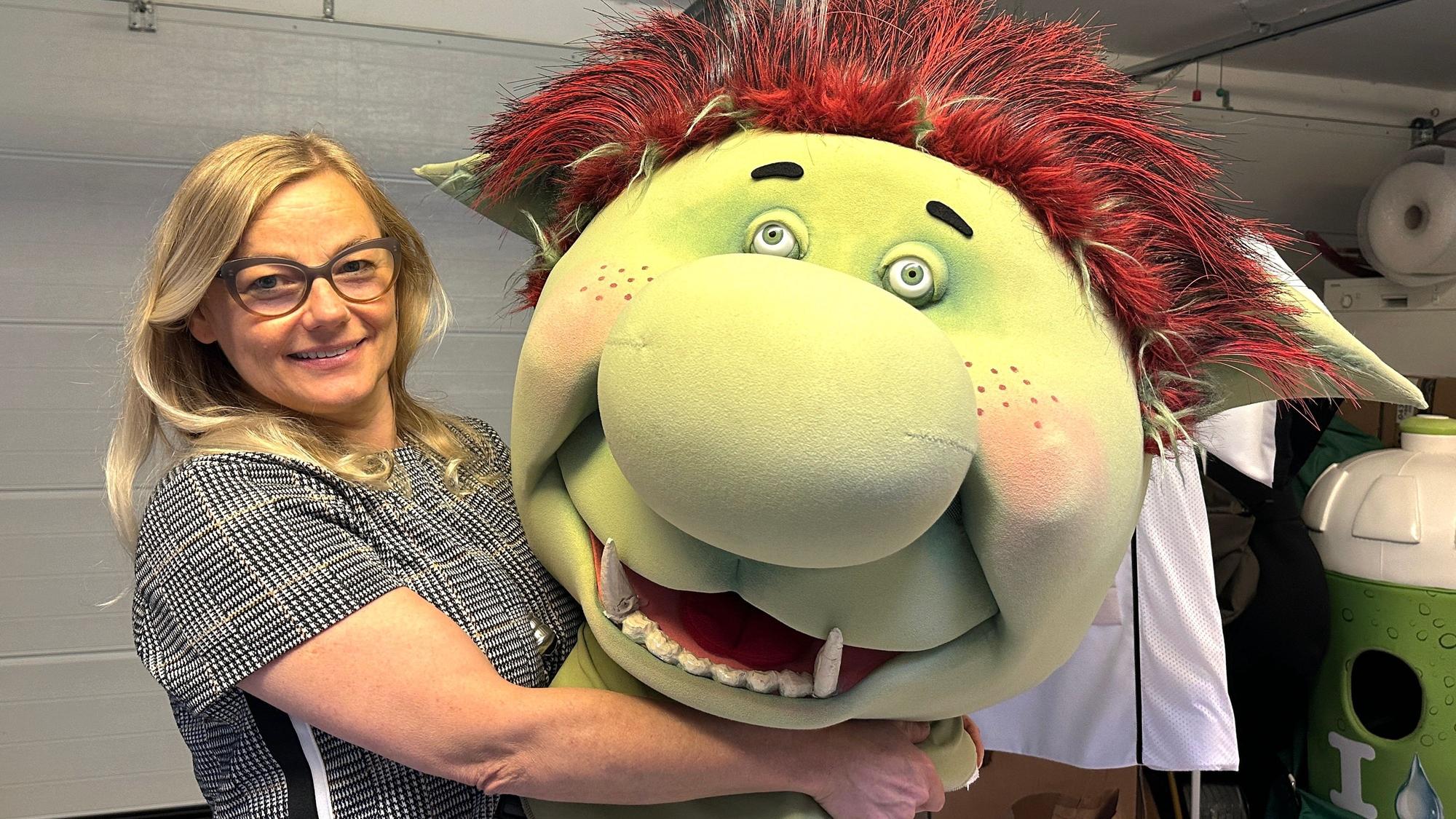 Eine Frau mit blondem Haar hat einen großen Stofftrollkopf im Arm.
