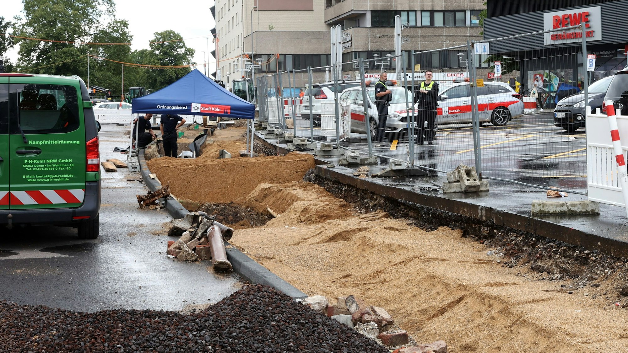 Blick auf eine Baustelle. In der Entfernung ist ein Zelt zu sehen, darunter stehen mehrere Personen. Rechts ist der Parkplatz eines Supermarktes sichtbar.