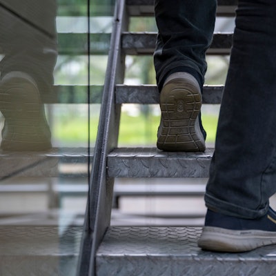 Ein Mann geht eine Treppe hoch. Es sind nur seine Füße zu sehen, die sich in dem Glasgeländer spiegeln.