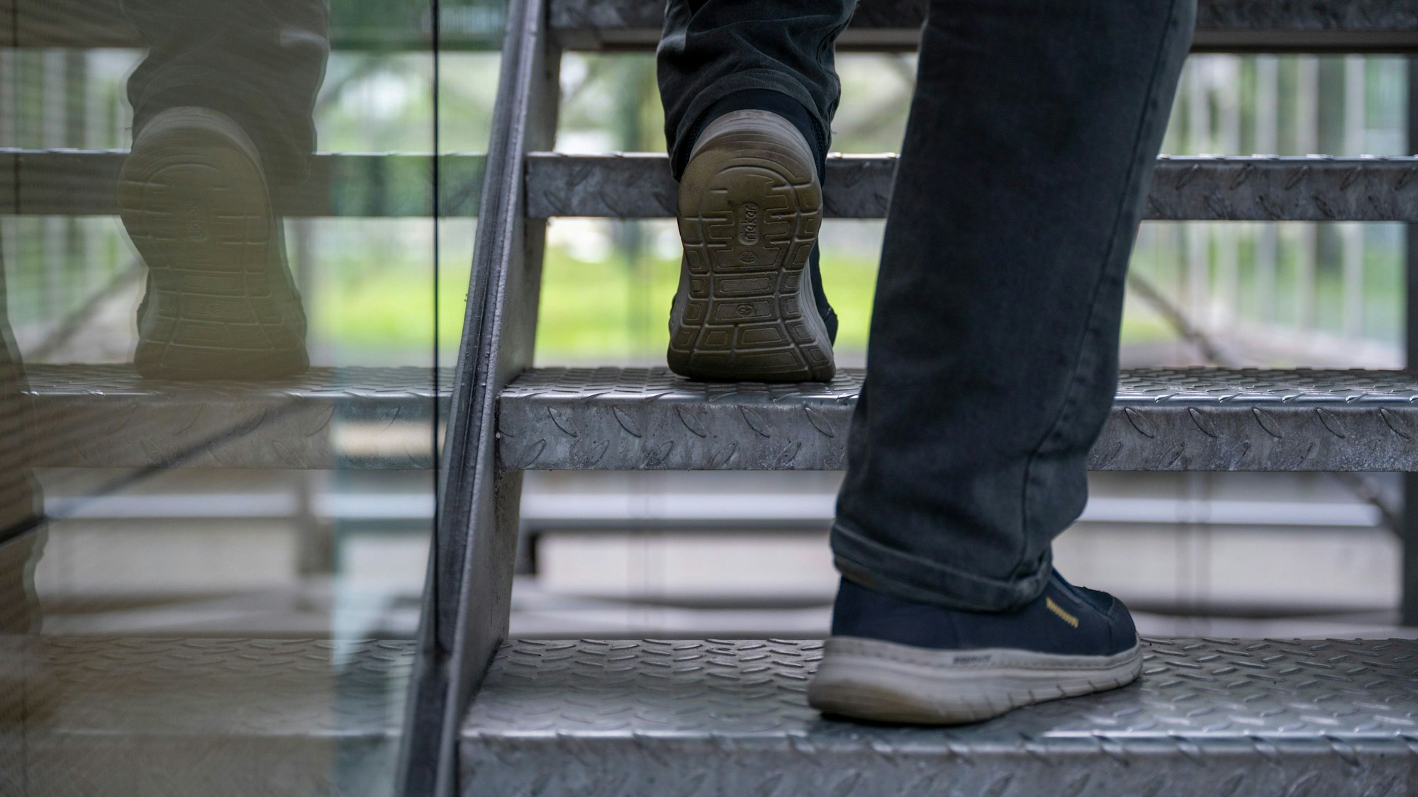 Ein Mann geht eine Treppe hoch. Es sind nur seine Füße zu sehen, die sich in dem Glasgeländer spiegeln.