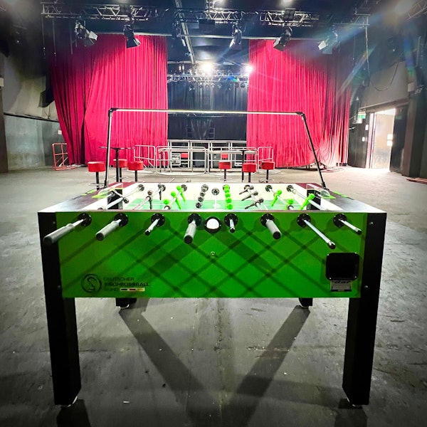 Ein Kickertisch steht in einem großen leeren Veranstaltungsraum, im Hintergrund ist eine Bühne mit einem roten Vorhang zu sehen.