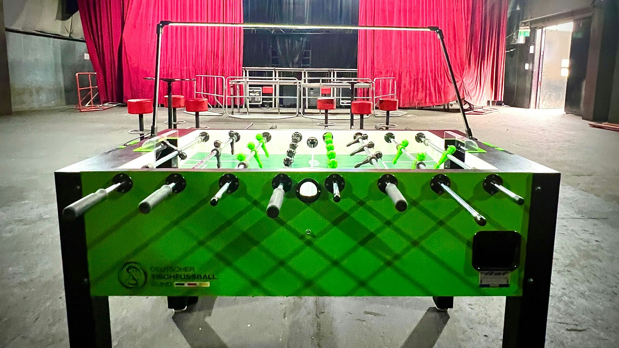 Ein Kickertisch steht in einem großen leeren Veranstaltungsraum, im Hintergrund ist eine Bühne mit einem roten Vorhang zu sehen.