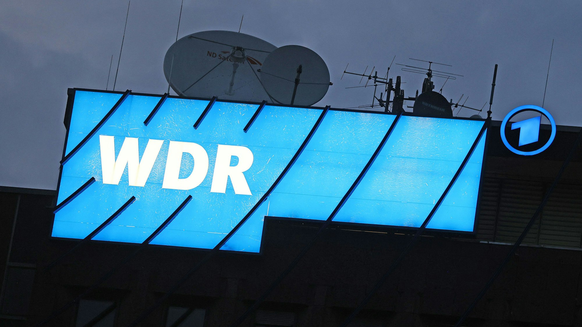 Das Logo des Nachrichtensenders WDR leuchtet auf dem Dach des WDR-Gebäudes in der Kölner Innenstadt.