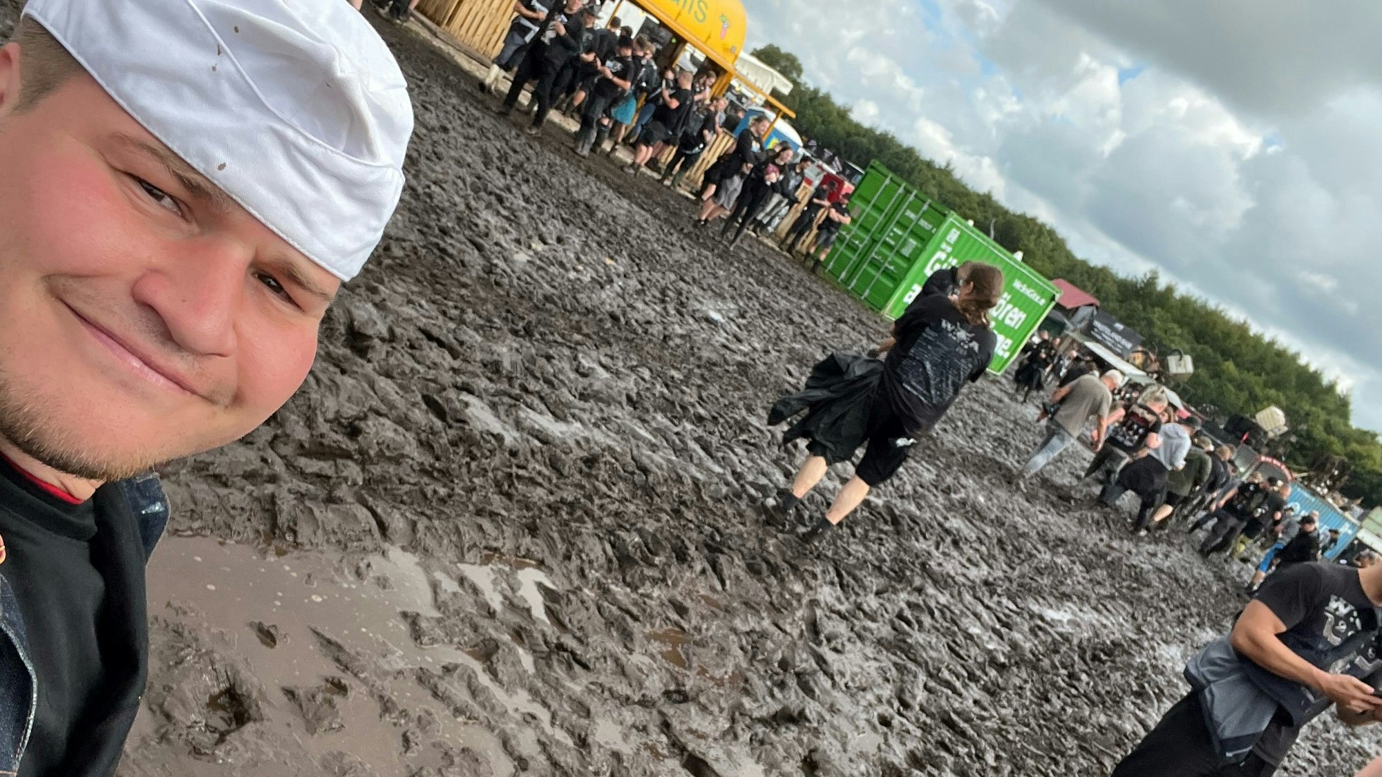 Das Selfie zeigt einen jungen Mann mit Bäckermütze und das schlammige Festival-Gelände von Wacken.