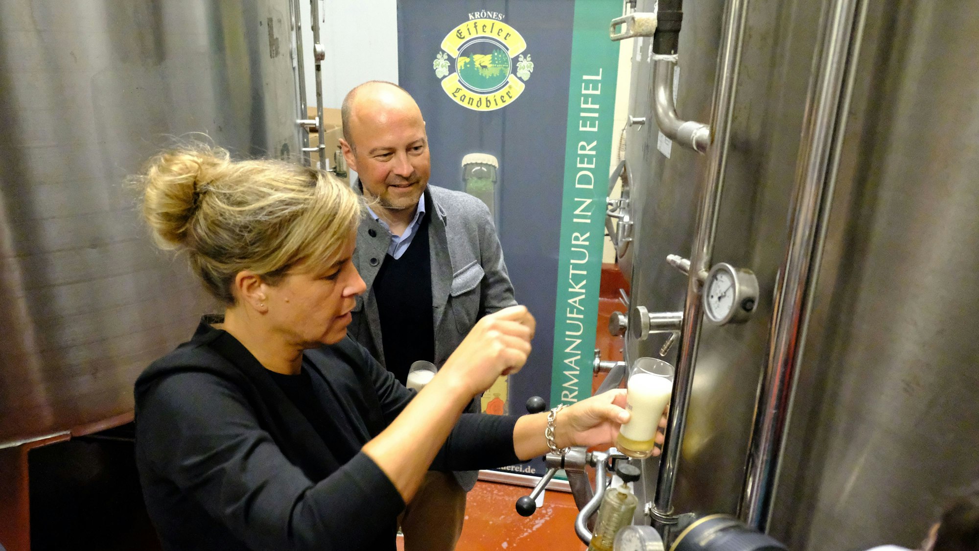 Eine Frau zapft ein Bier aus einem silberfarbenen Stahltank einer Brauerei. Eiin Mann beobachtet sie dabei.