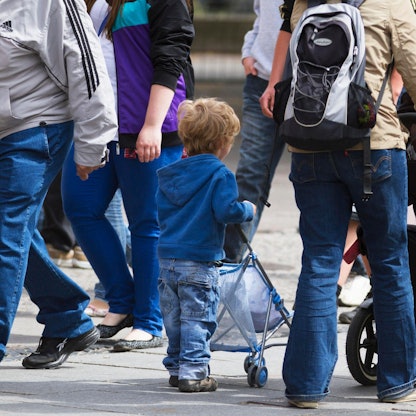 Ein kleiner Junge schiebt einen Puppenwagen, neben ihm läuft seine Mutter mit einem Kinderwagen.
