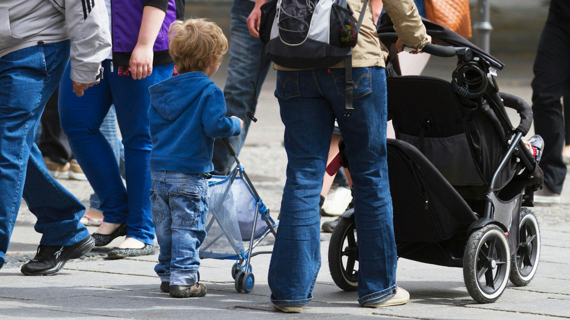 Ein kleiner Junge schiebt einen Puppenwagen, neben ihm läuft seine Mutter mit einem Kinderwagen.