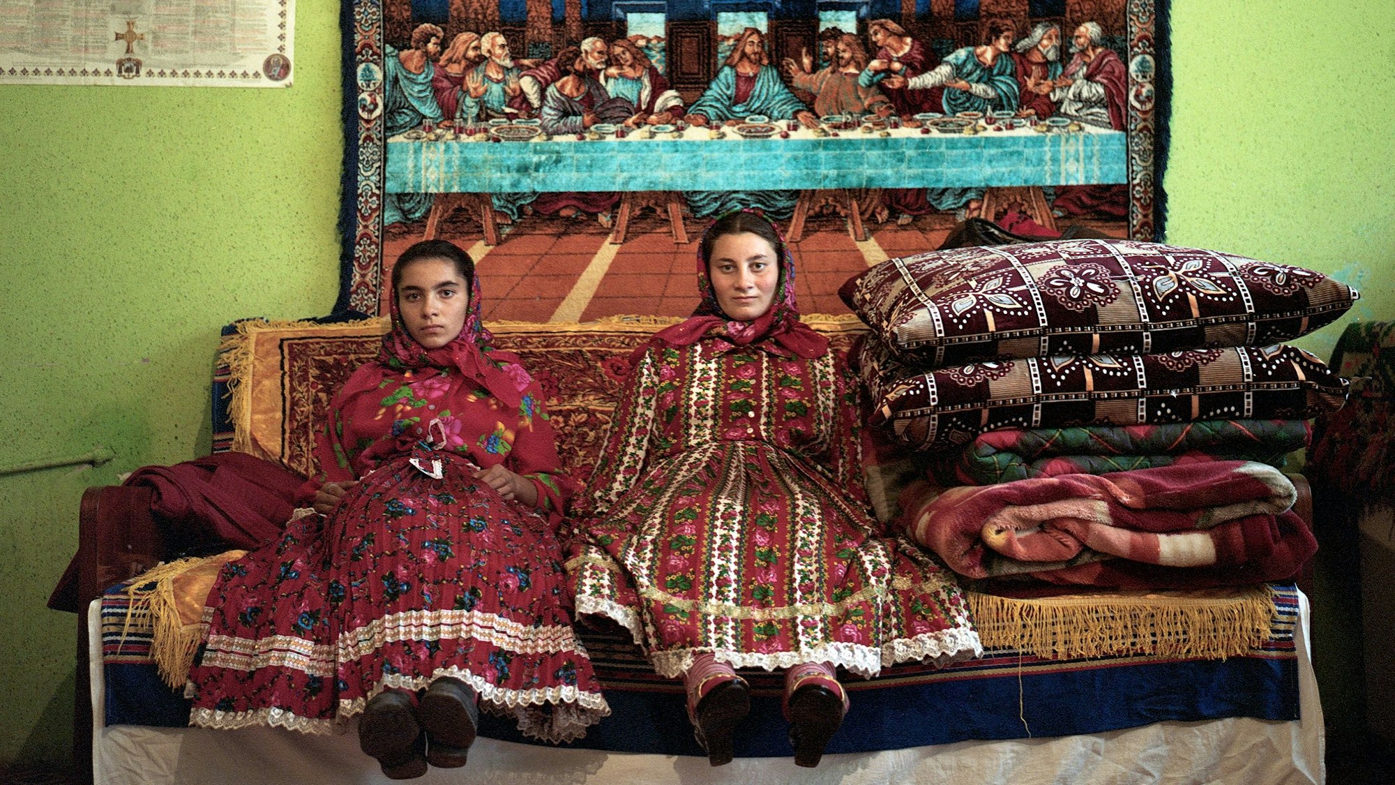 Zwei Frauen in traditioneller Kleidung vor einem Wandteppich, auf einer großen Couch.