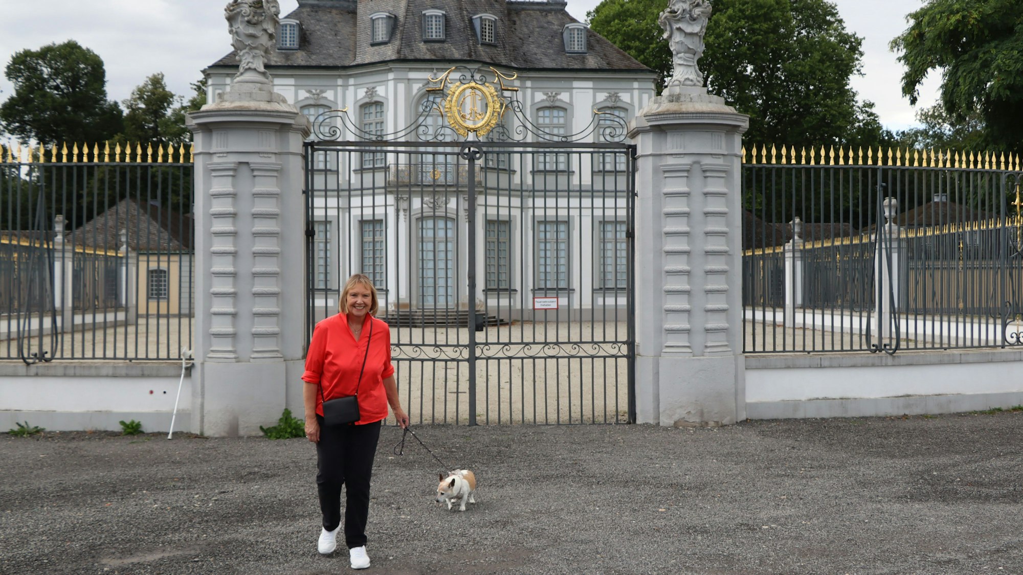 Auf dem Foto ist eine Spaziergängerin mit ihrem Hund vor einem schmiedeeisernen Tor und einem Schloss im Hintergrund zu sehen.