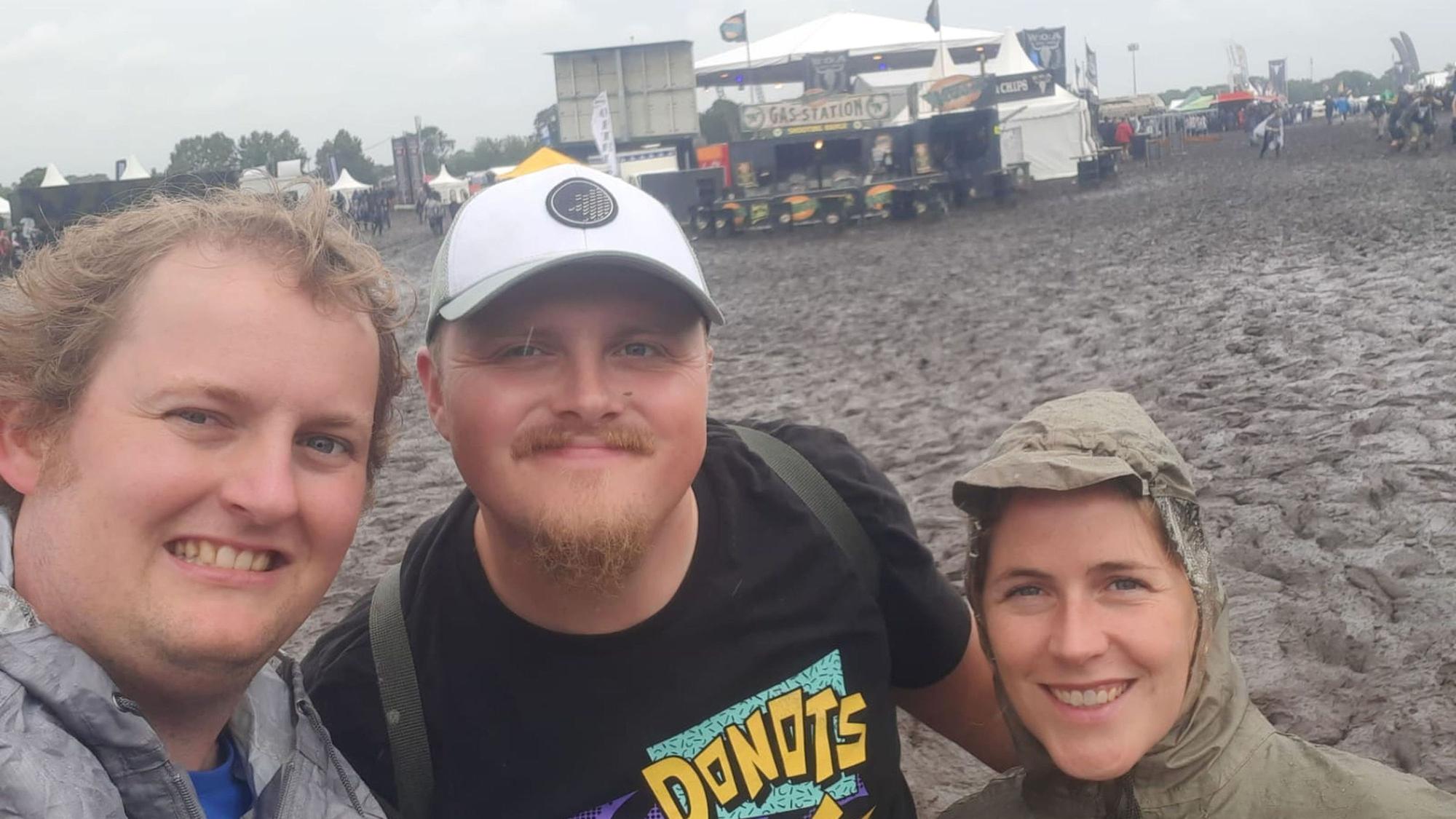 Drei Besucher des Festivals vor dem schlammigen Boden an einer Bühne.