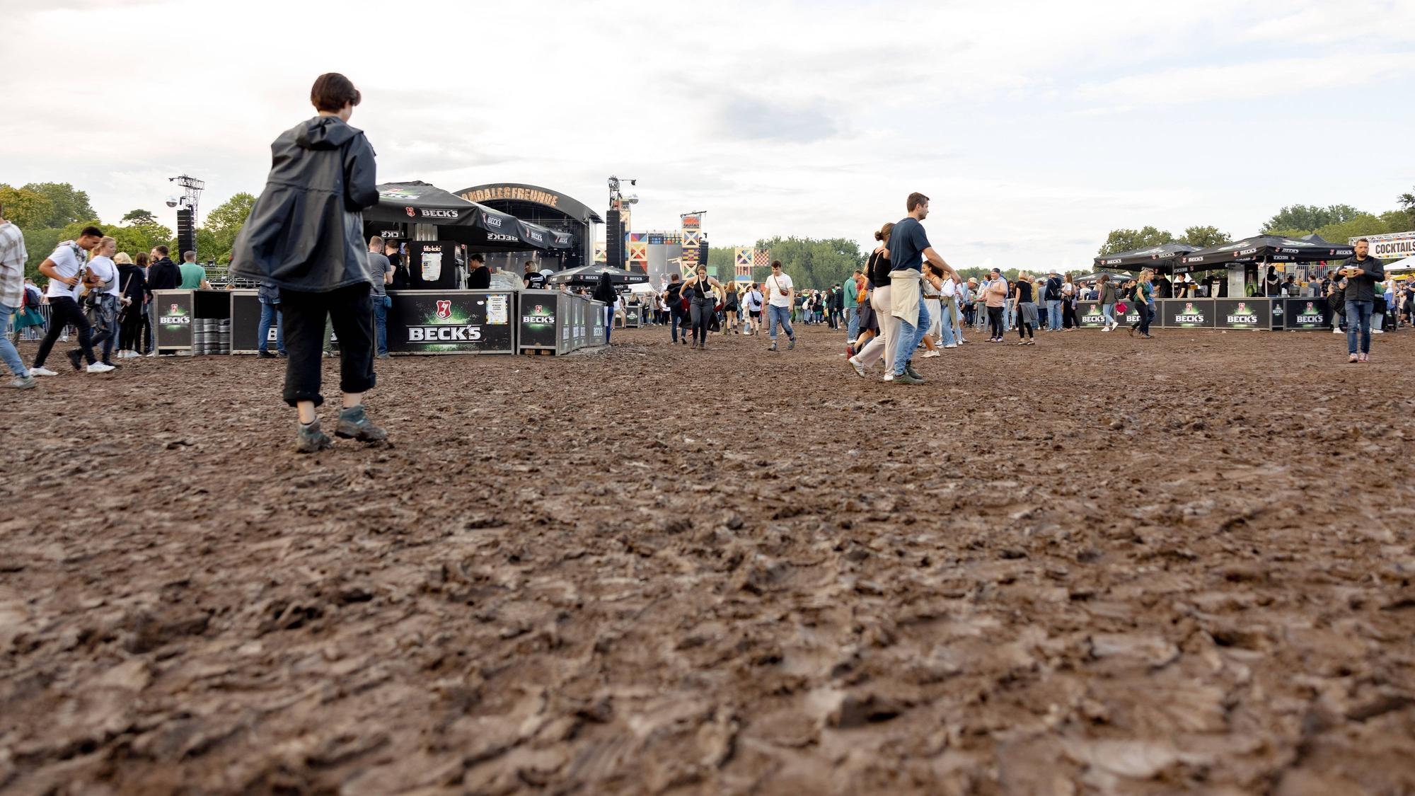 Festivalbesucher gehen in der Bonner Rheinaue vor den Bühnen über eine große Matschfläche. Es ist bewölkt.