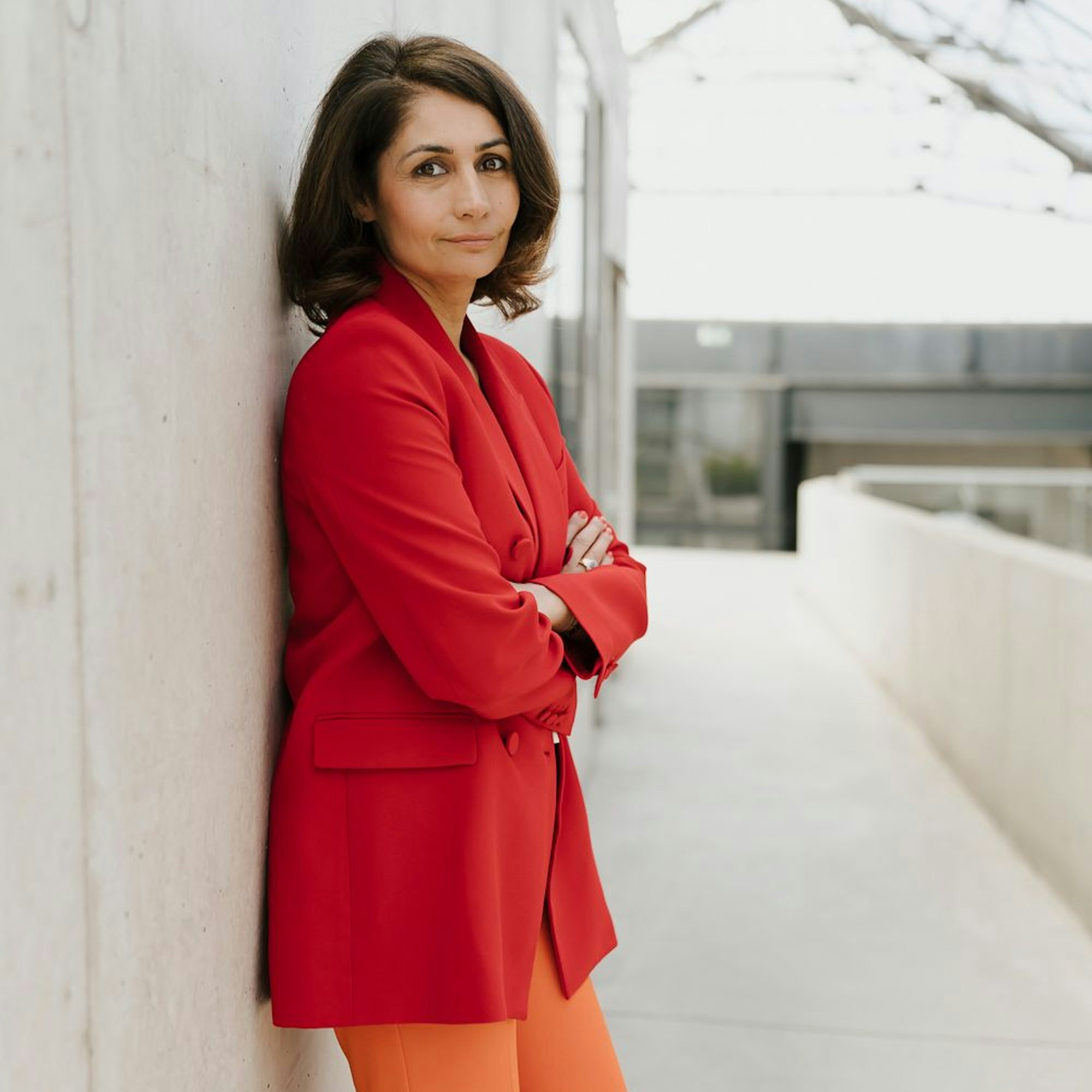 Leila Cheraghi im roten Blazer an eine Wand gelehnt.