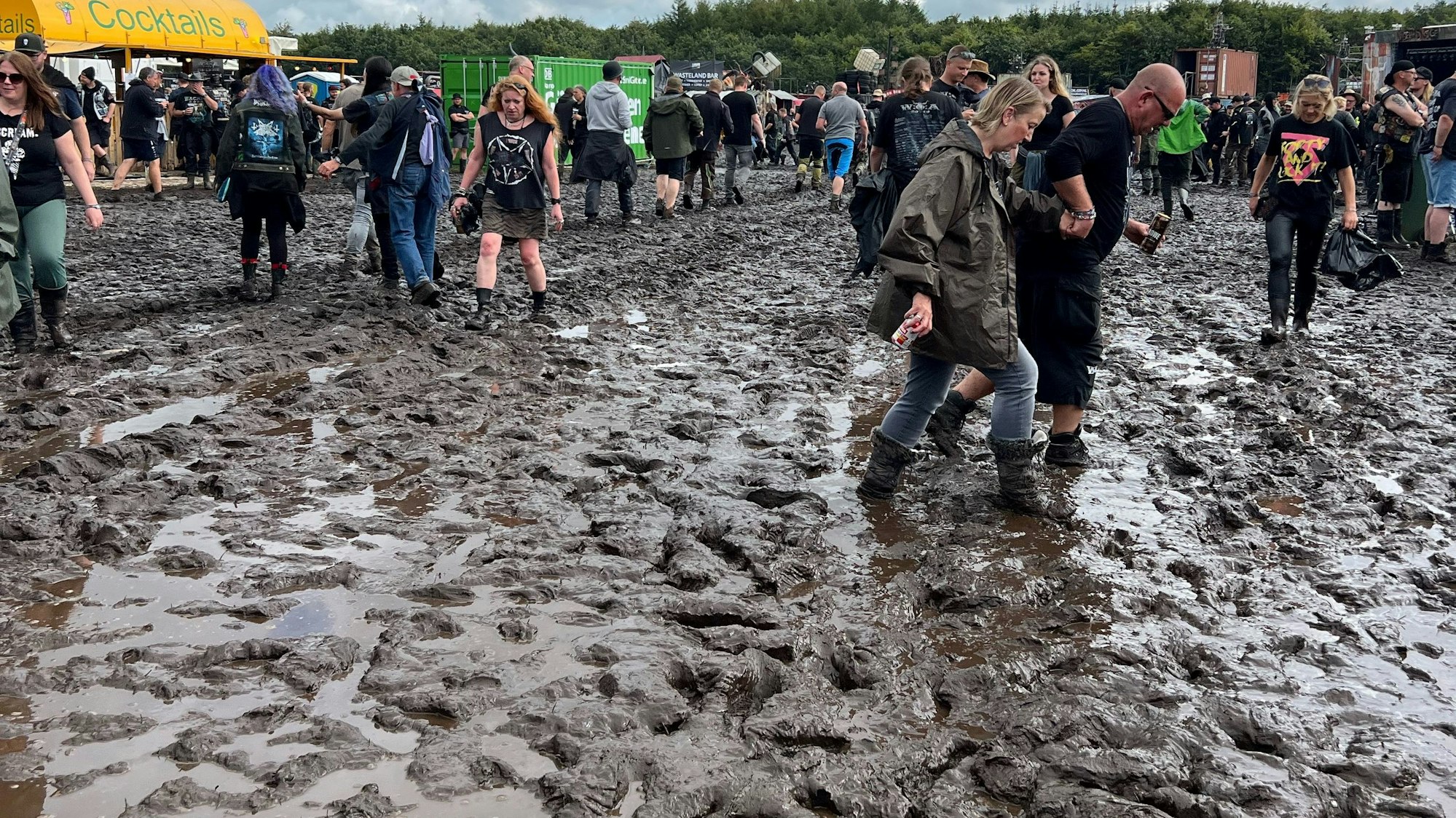 Das Bild zeigt das Festival-Gelände von Wacken und einige Fans, die in Gummistiefeln durch tiefen Schlamm.