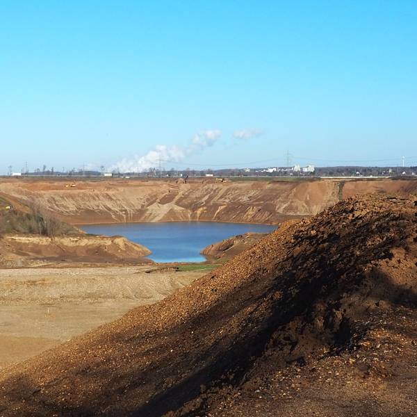 Hinter Hügeln aus braunem Sand und Kies ist eine große Fläche Wasser zu sehen, das sich in der Kiesgrube ansammelt. Links am Rand der Grube arbeiten Bagger.