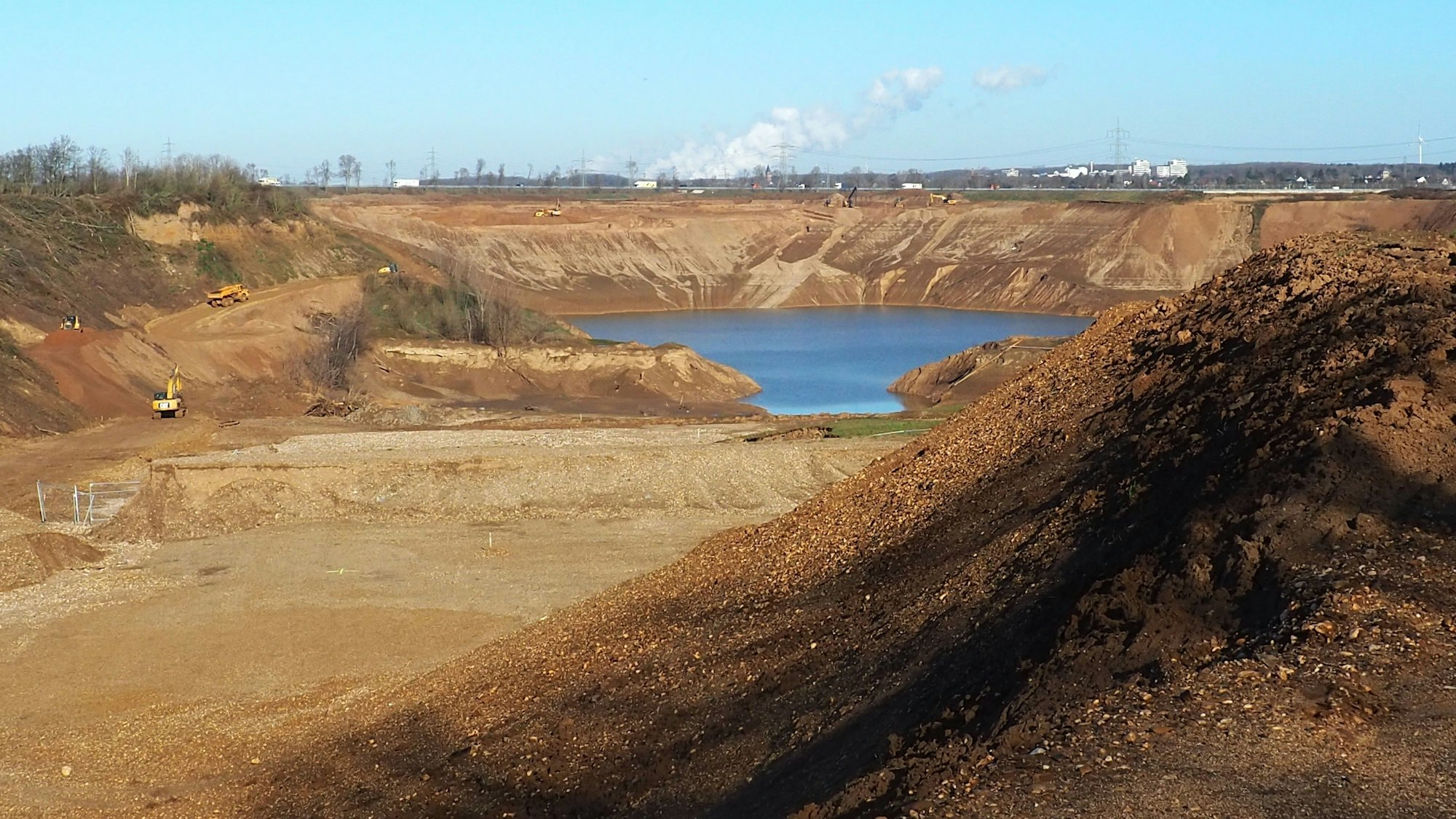 Hinter Hügeln aus braunem Sand und Kies ist eine große Fläche Wasser zu sehen, das sich in der Kiesgrube ansammelt. Links am Rand der Grube arbeiten Bagger.