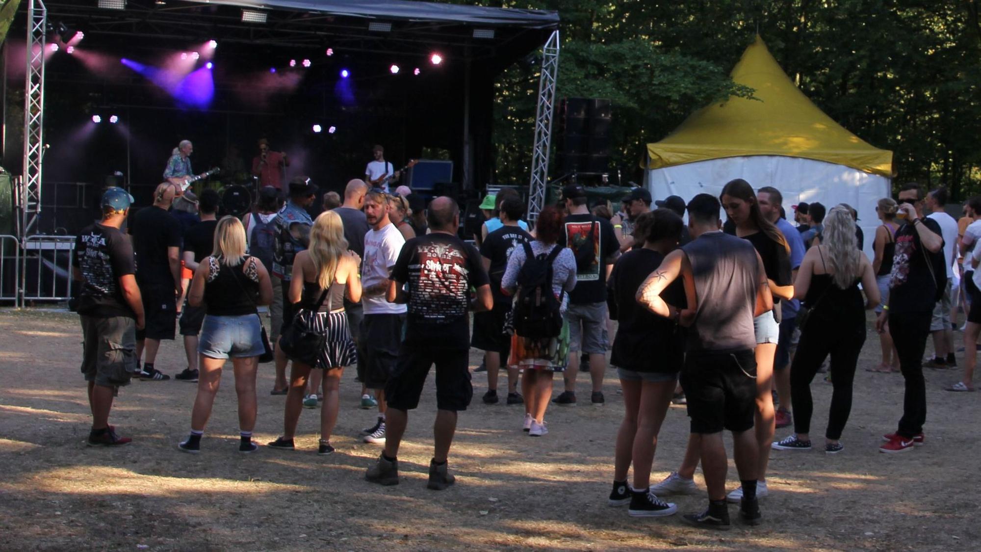 Vor einer kleinen Bühne im Wald stehen Fans und hören der Band zu.