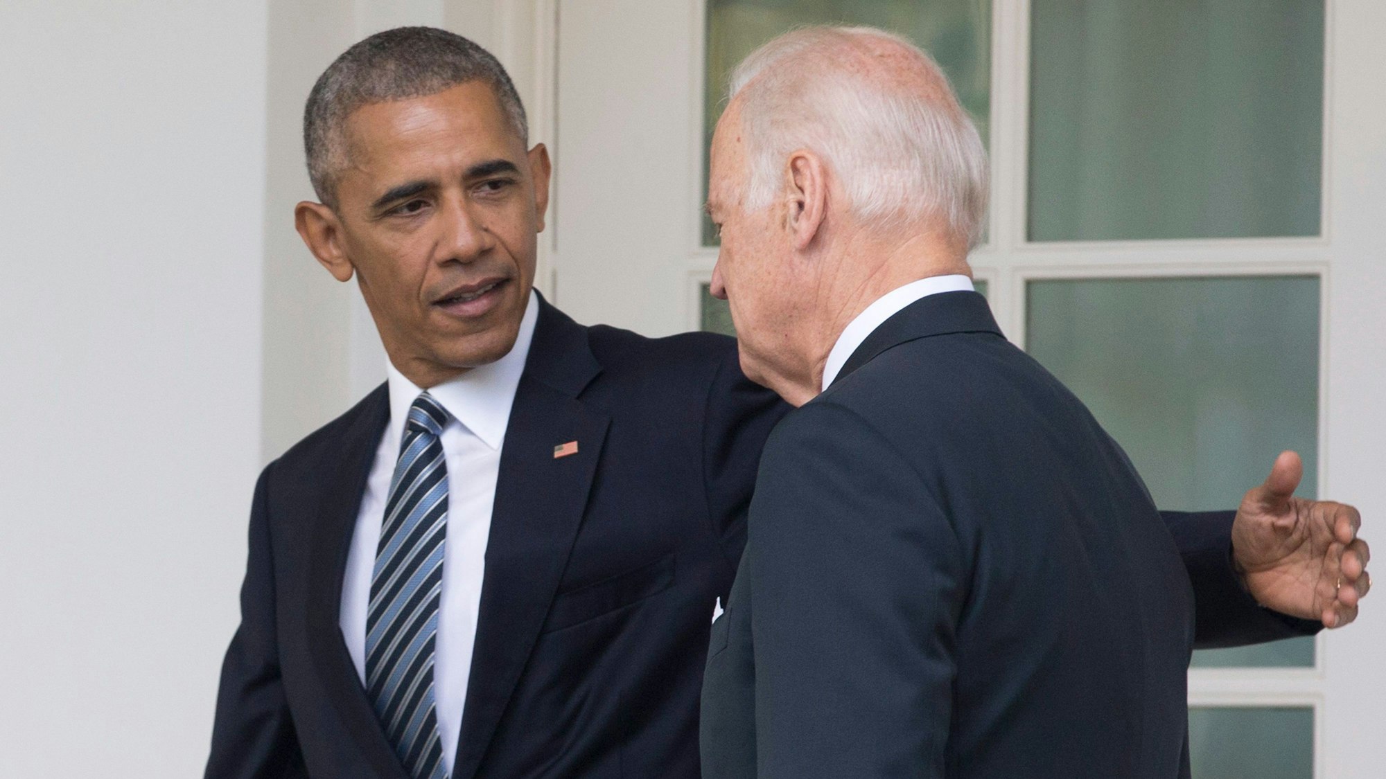 Barack Obama (l.) schaut Joe Biden (r.) an und hält seinen Arm hinter den Rücken des US-Präsidenten. Beide Männer tragen einen Anzug.