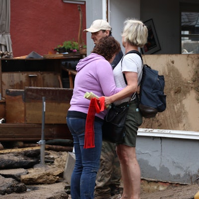 Das Bild zeigt zwei Frauen, die sich nach der Flutkatastrophe in Bad Münstereifel trösten. Im Hintergrund sind ein Mann und ein zerstörtes Haus zu sehen.