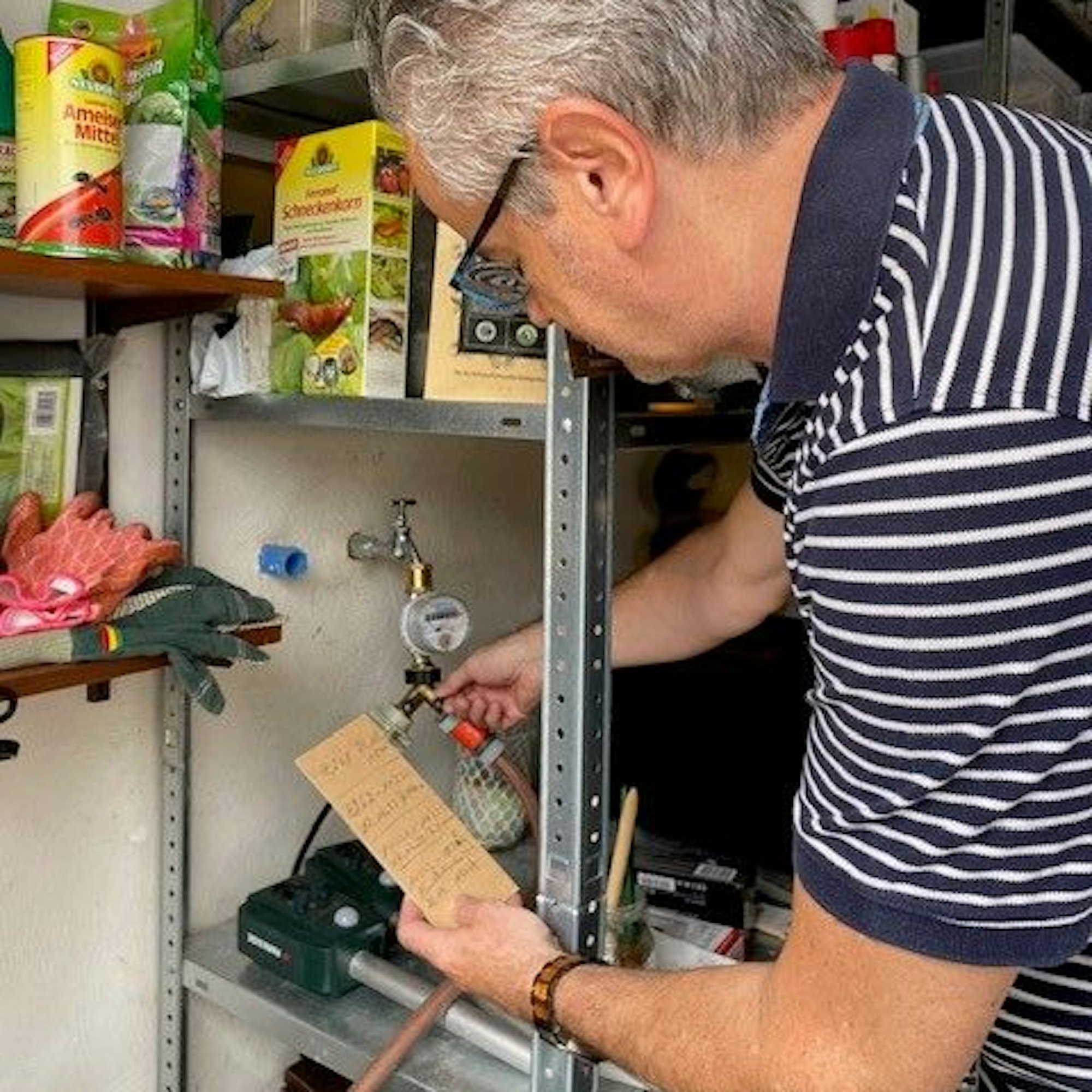 Addi Kirfel dreht mit einer Hand an der Wasserzufuhr, mit der anderen hält er eine kleine Pappe, auf der er mit Bleistift den Wasserverbrauch notiert.