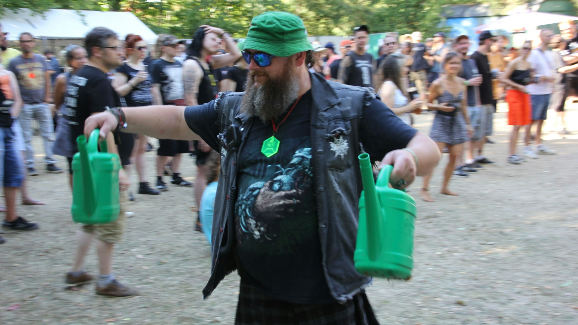 Ein Metal-Fan mit grünem Hut trägt zwei grüne Gießkannen durch das Publikum.