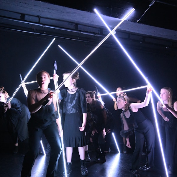 Jugendliche stehen auf der Bühne und halten sich an leuchtenden Stäben, die wie ein Haus aufgebaut sind, fest.
