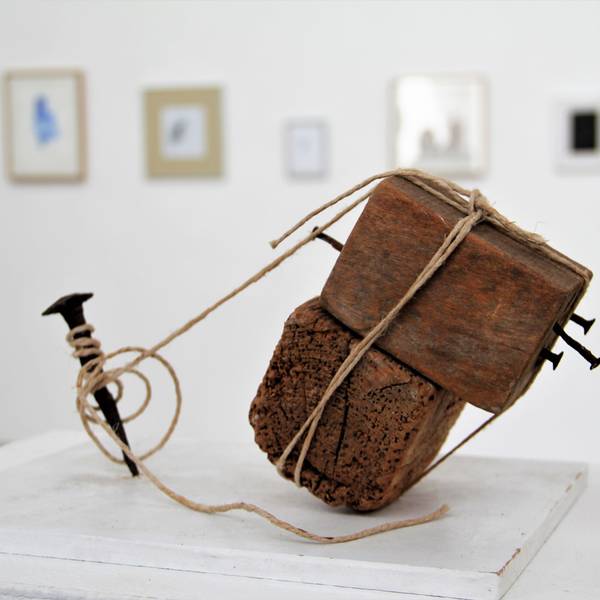 Ein Holzobjekt, das mit Bindfäden an einem Nagel befestigt ist, steht in einem Ausstellungsraum.
