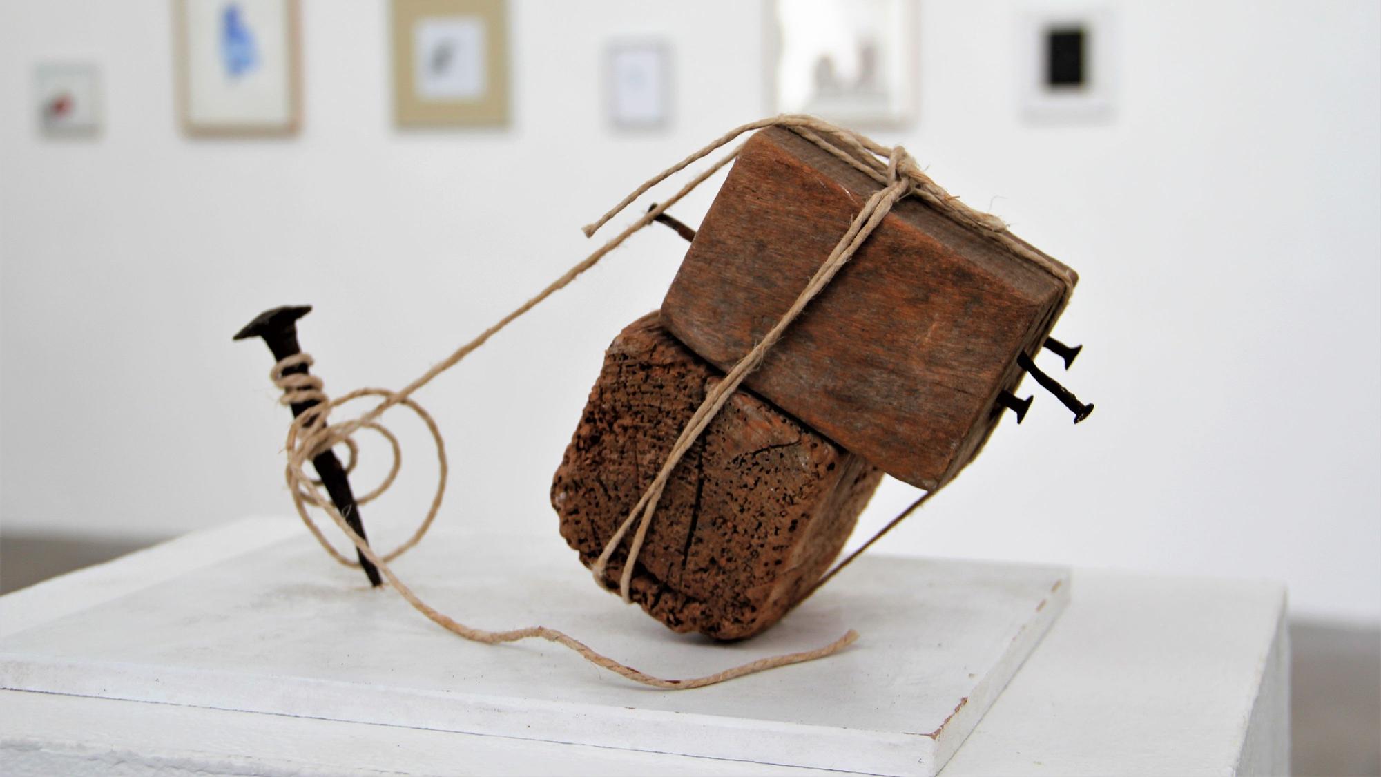 Ein Holzobjekt, das mit Bindfäden an einem Nagel befestigt ist, steht in einem Ausstellungsraum.