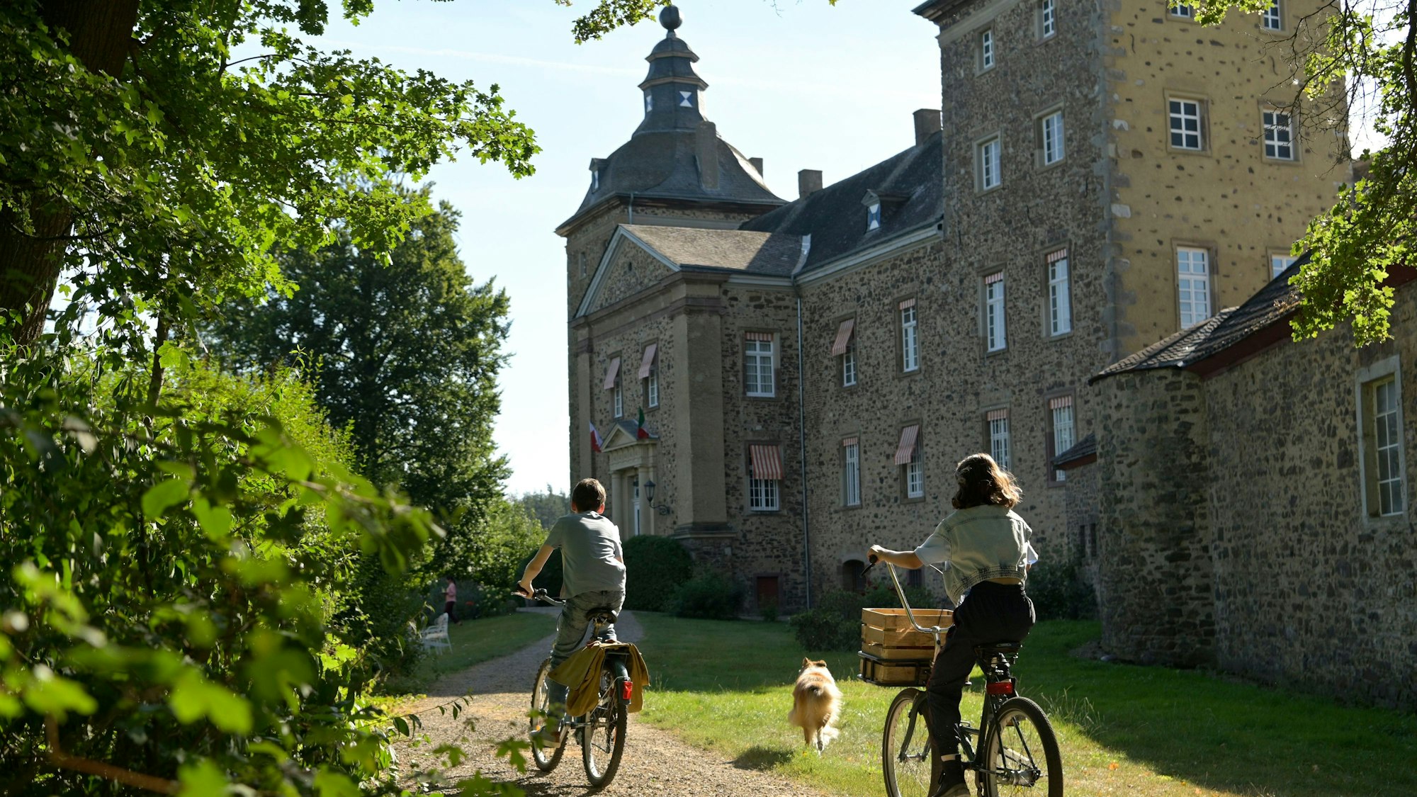 Auf dem Bild sind zwei Schauspieler zu sehen, die mit dem Fahrrad an der Burg Ringsheim vorbeifahren. Auch Lassie ist zu sehen.