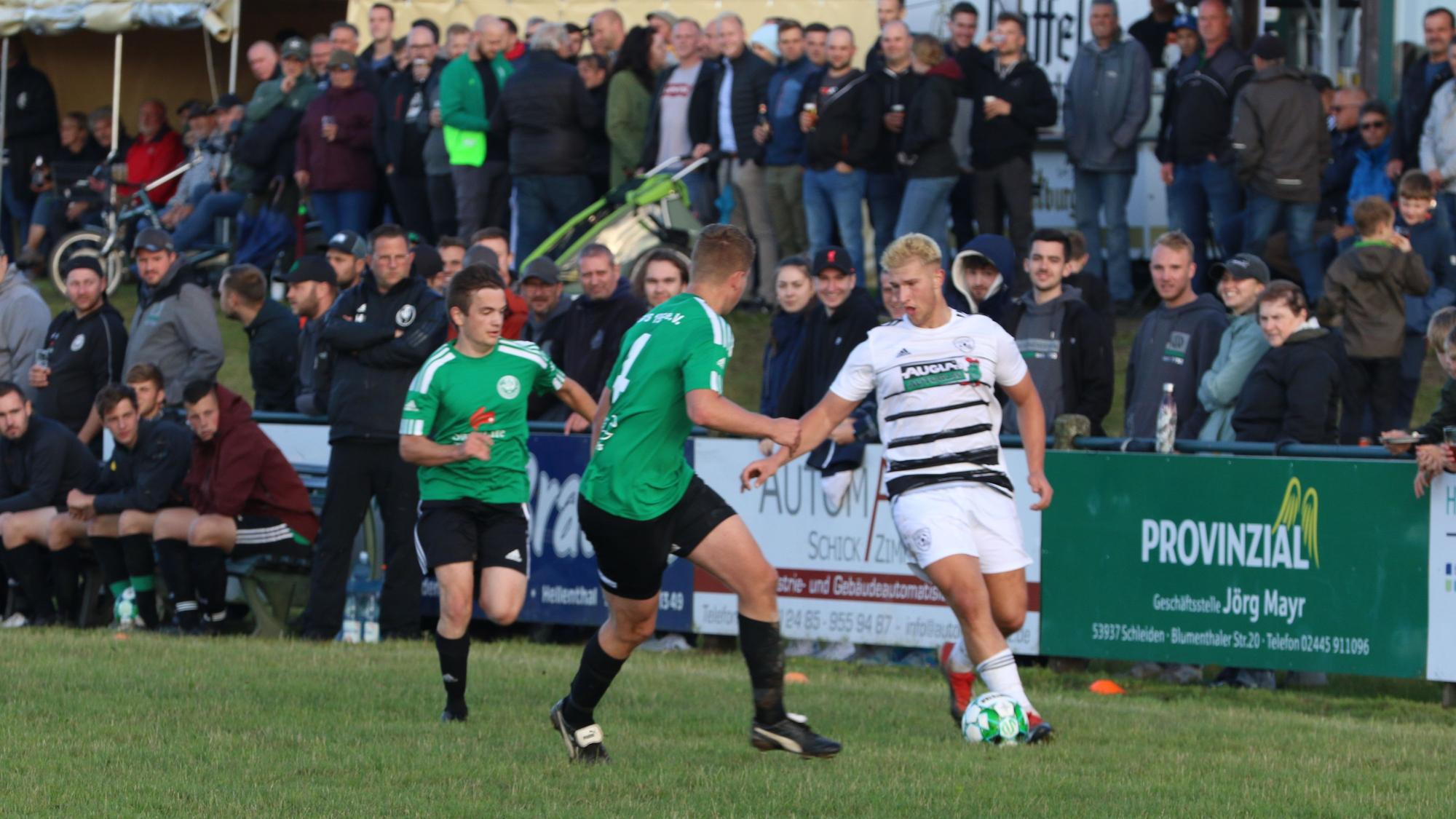 Ein Erftstädter Spieler kontrolliert, bedrängt von zwei Spielern aus Schöneseiffen, den Ball. Im Hintergrund sind die Zuschauer zu sehen.