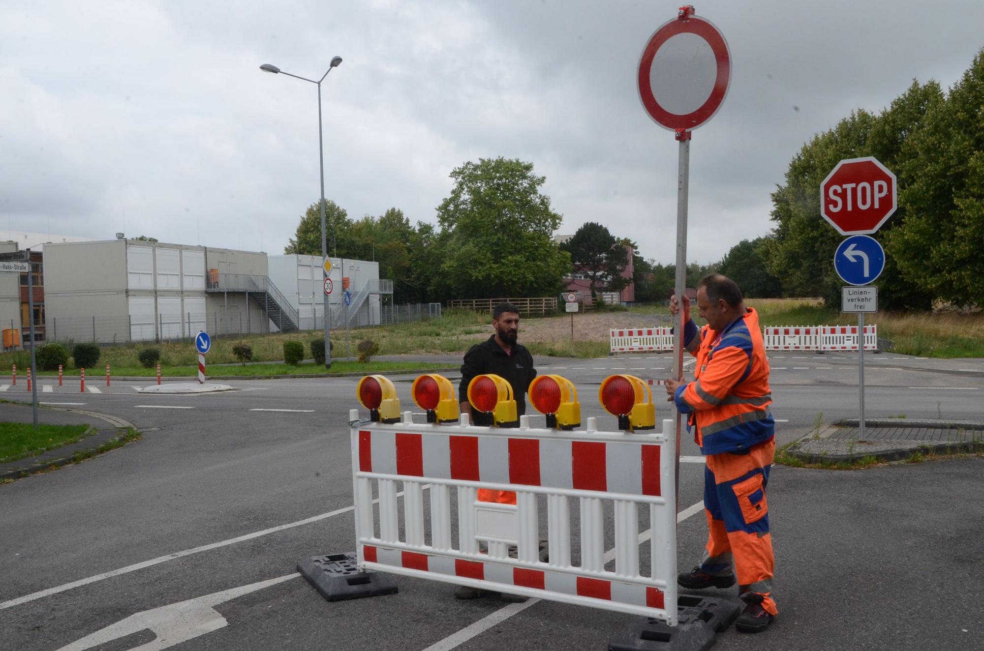 Zwei Männer stellen Absperrungen und Verkehrsschilder auf, einer trägt eine orange Jacke. Im Hintergrund stehen Container als Schulräume.