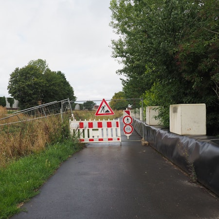 Ein asphaltierter Weg ist mit Baken und Bauzaun-Elementen versperrt.